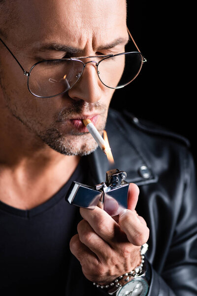 Handsome man in sunglasses lighting cigarette with lighter isolated on black