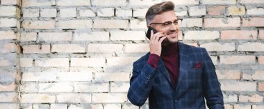 Arka planda tuğla duvarla akıllı telefonla konuşan gülümseyen iş adamının panoramik görüntüsü.