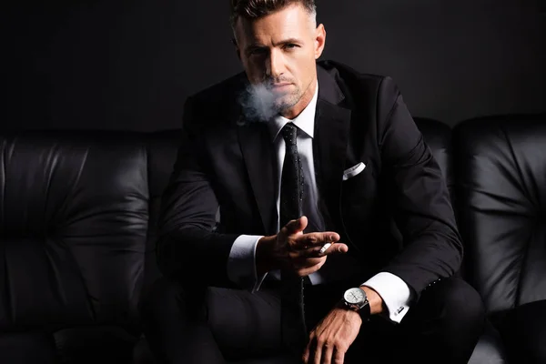 黒で隔離されたソファの上でハンサムなビジネスマンの喫煙タバコ — ストック写真