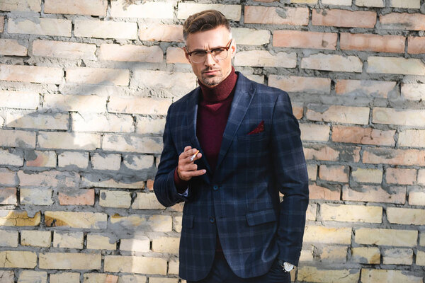 Красивый бизнесмен держит сигарету и смотрит в камеру с кирпичной стеной на заднем плане
