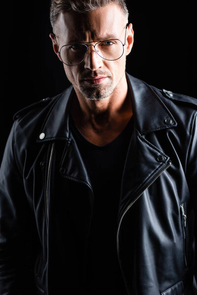 Serious man in biker jacket and sunglasses looking at camera isolated on black