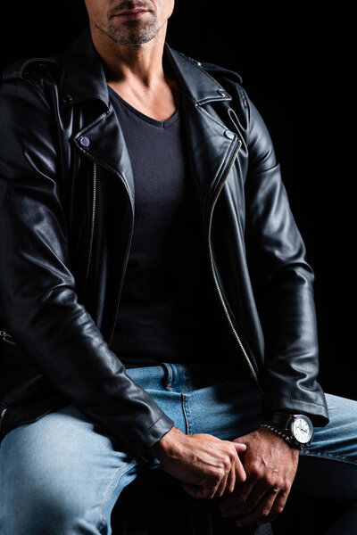 Cropped view of stylish man in leather jacket and jeans sitting on chair isolated on black 