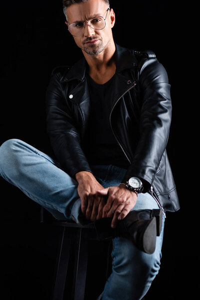 Confident man in biker jacket and jeans looking at camera while sitting on chair isolated on black 