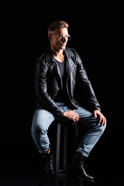 Full length of stylish man in jeans and leather jacket sitting on chair isolated on black