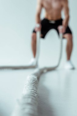 Çıplak gövdeli seksi kaslı vücut geliştiricinin seçici odağı gri arka planda savaş halatıyla egzersiz yapıyor.