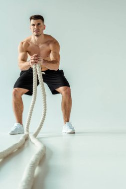 Çıplak gövdeli seksi kaslı vücut geliştiricinin seçici odağı gri arka planda savaş halatıyla egzersiz yapıyor.
