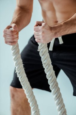 Çıplak gövde egzersizi yapan seksi kaslı vücutçunun gri üzerinde izole edilmiş savaş halatıyla görüntüsü.