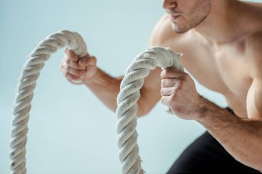 Çıplak gövde egzersizi yapan seksi kaslı vücutçunun gri üzerinde izole edilmiş savaş halatıyla görüntüsü.