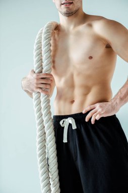 Çıplak gövdeli seksi kaslı vücut geliştirici, gri üzerinde izole edilmiş savaş halatıyla poz veriyor.