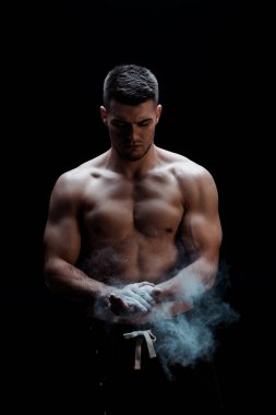 Çıplak gövdeli seksi kaslı vücut geliştirici kara arka planda dumanlı talk pudrası uyguluyor. 
