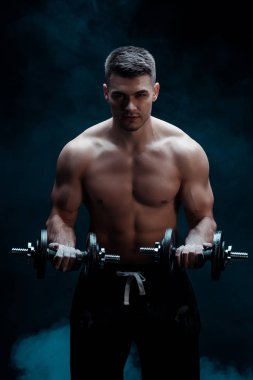 Çıplak gövdeli seksi kaslı vücut geliştirici. Dumanla siyah halterleri kesiyor.