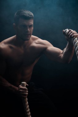 Çıplak gövdeli seksi kaslı vücut geliştirici. Siyah arka planda savaş halatıyla kesilen dumanlı. 