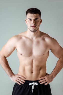 Çıplak gövdeli seksi kaslı vücut geliştirici elleri kalçasında gri renkte poz veriyor.