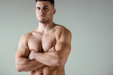 Çıplak vücutlu seksi kaslı vücut geliştirici kollarını çapraz yapıp gri renkte poz veriyor.