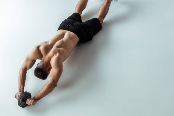 overhead view of sexy muscular bodybuilder with bare torso exercising with ab wheel on grey background
