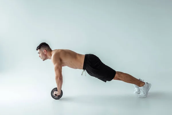 Side view of sexy muscular bodybuilder with bare torso exercising with ab wheel on grey background — Stock Image Side View Sexy Muscular Bodybuilder Bare Torso Exercising Wheel Grey Royalty Free Stock Photos