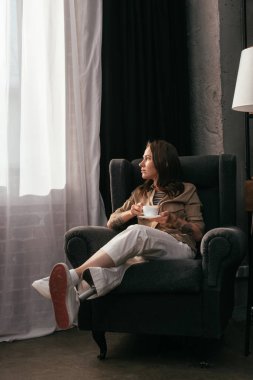 Elinde kahve fincanı tutan ve oturma odasındaki pencereye bakan bacak protezi olan kız.
