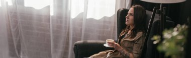 Panoramic shot of beautiful woman holding coffee cup while sitting in armchair at home clipart