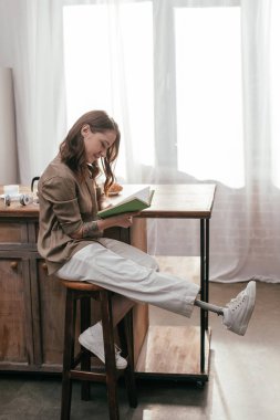 Gülümseyen bir kadının bacak protezi ile evde masa başında kitap okuması.