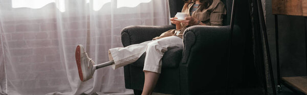 Обрезанный вид девушки с протезной ногой, держащей чашку кофе, сидя в кресле, панорамный снимок
