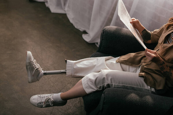 Обрезанный вид женщины с протезной ногой читающей газету в кресле
