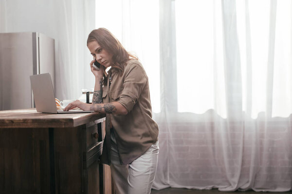 Молодая женщина разговаривает на смартфоне и с помощью ноутбука на кухонном столе
