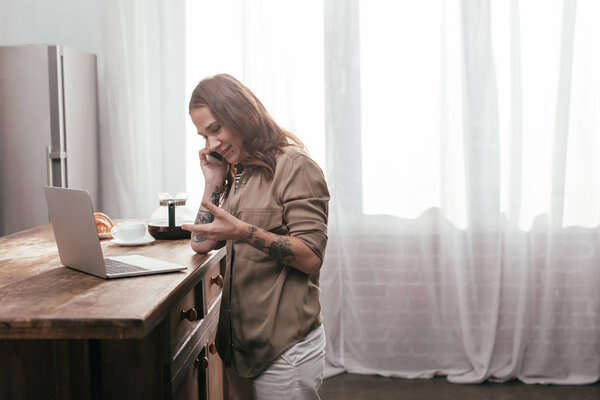 Вид сбоку улыбающейся женщины, говорящей по смартфону рядом с ноутбуком на кухонном столе
