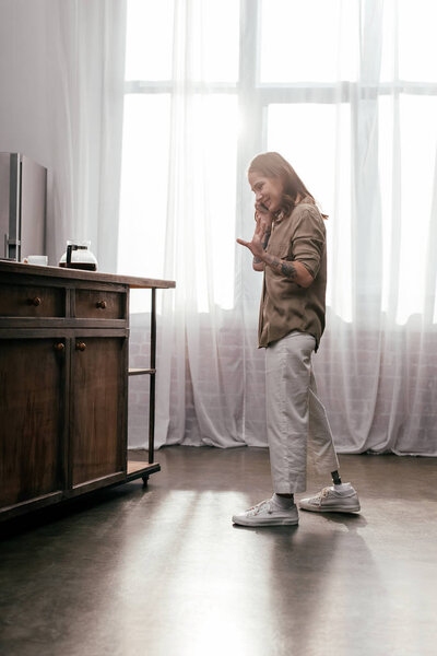 Вид сбоку улыбающейся женщины с протезом ноги, говорящей по смартфону на кухне
 