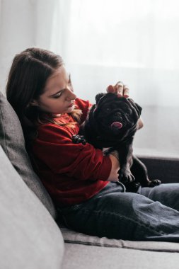 Kanepede köpeğini okşayan genç bir kadının yan görüntüsü.