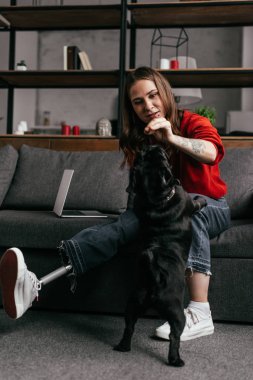 Protez bacaklı genç bir kadın oturma odasında köpeğini besliyor.