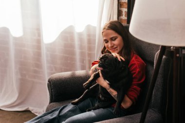 Protez bacaklı neşeli kadın evdeki koltukta köpeğini okşuyor.