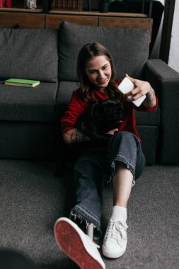 Protez bacaklı bir kız köpeğini tutuyor ve oturma odasında yerde akıllı telefonuyla selfie çekiyor.
