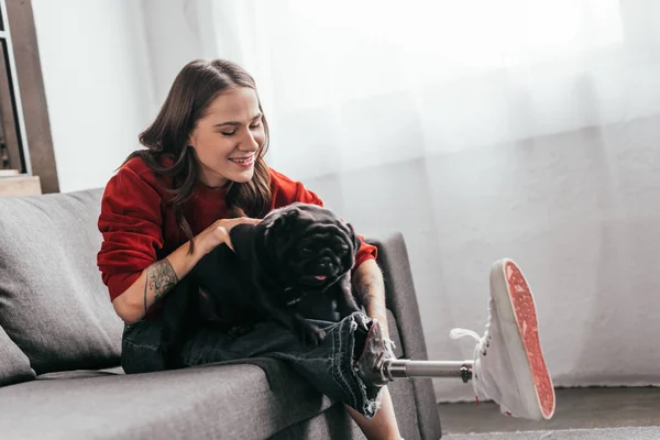 Lächelnde Frau Mit Beinprothese Spielt Mit Mops Auf Sofa — Stockfoto