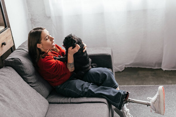 Вид сбоку женщины с протезной ногой, играющей с мопсом на диване
