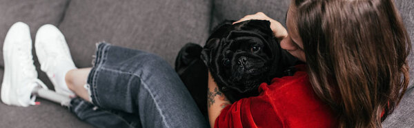 Selective focus of girl with leg prosthesis petting pug on sofa, panoramic shot