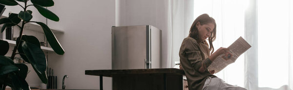 Side view of woman with leg prosthesis reading newspaper at kitchen, panoramic shot