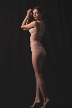 İç çamaşırlı, siyah arka planda poz verirken kameraya bakan çekici genç bir kadının tam boy görüntüsü.