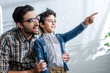 Gülümseyen yahudi oğlu parmağıyla işaret ediyor ve babası evdeyken pencereden bakıyor. 