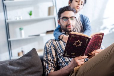 selective focus of jewish father and son reading tanakh in apartment  clipart