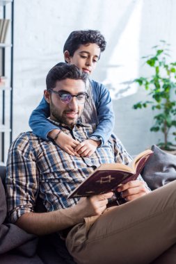 smiling jewish father and son reading tanakh in apartment  clipart