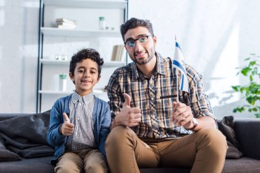 smiling jewish father with flag of israel and son showing thumbs up in apartment  clipart
