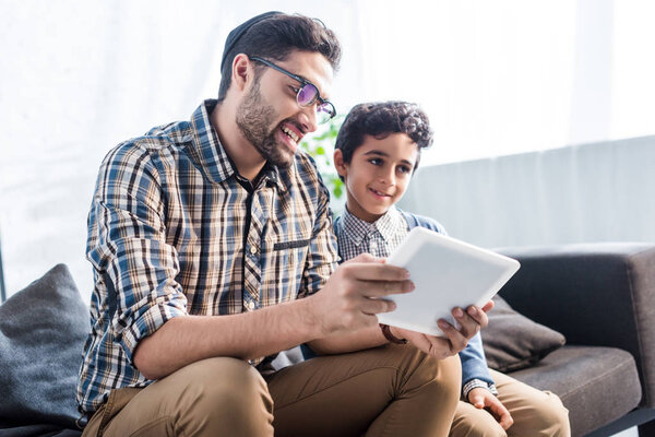smiling jewish father and son using digital tablet in apartment 