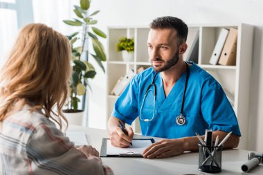 selective focus of bearded doctor holding pen and looking at woman  clipart