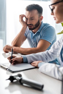 selective focus of bearded man looking at doctor in glasses writing diagnosis 