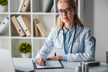 attractive doctor in glasses writing diagnosis near laptop 