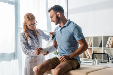 attractive orthopedist putting bandage on injured arm of bearded man 