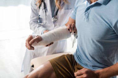 cropped view of orthopedist putting bandage on injured hand of man 