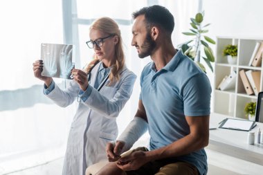 attractive orthopedist in glasses holding x-ray near handsome injured man 
