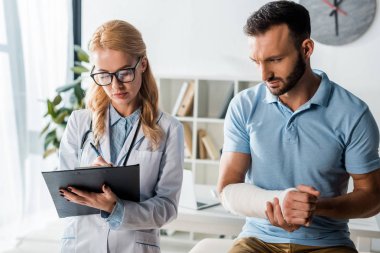 attractive orthopedist holding clipboard near injured bearded man 