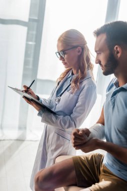 attractive orthopedist writing diagnosis near injured bearded man 